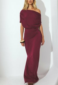 Kvinna iklädd en lång, avskuren burgundy-klänning med rynkade ärmar och två bangles på sin vänstra handled mot en vit bakgrund.