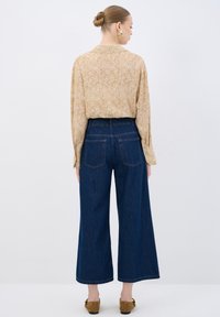 Blusa beige con estampado floral y mangas largas; combinada con pantalones de mezclilla de pierna ancha en azul oscuro. Zapatos de ante marrón visibles en la parte inferior.