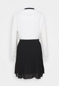 Blouse blanche à manches longues avec col à volants et boutons dorés, associée à une jupe plissée noire taille haute, vue de dos.