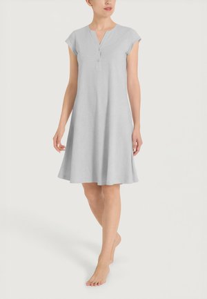 Donna che indossa un vestito grigio chiaro, lungo fino al ginocchio, a maniche corte con bottoni allo scollo, in piedi a piedi nudi su uno sfondo semplice.