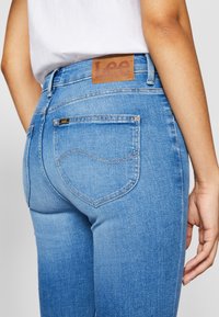 Blue denim jeans with a high waist, featuring a brown leather patch labeled "Lee" and curved back pockets with stitching details.