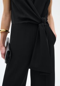 Jumper noir avec un design avant torsadé et une ceinture nouée à la taille. Les accessoires comprennent une pochette noire élégante et un bracelet en or.