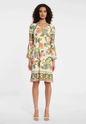 Frau trägt ein knieumspieltes cremefarbenes Kleid mit bunten Blumenmustern und ausgestellten Ärmeln, kombiniert mit beigen Ankle-Strap-Pumps.