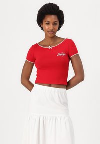 Top corto rojo de manga corta con un escote amplio y ribete blanco. Presenta un pequeño lazo blanco y un logo impreso en forma de corazón.