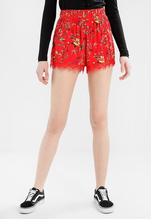Mujer con pantalones cortos rojos con encaje floral, camiseta negra de manga larga y zapatillas negras y blancas, de pie contra un fondo blanco.