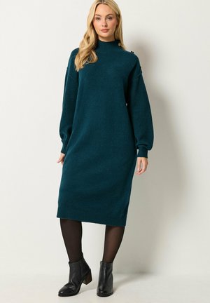 Femme aux longs cheveux blonds portant une robe midi en maille à manches longues couleur sarcelle, des collants noirs et des bottines noires, debout devant un fond uni.