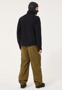 Black zip-up jacket, olive-green baggy pants, knitted mask, textured fabric, and visible stitching details. Black footwear completes the outfit.