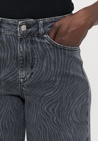 Črne denim kavbojke z diskretno valovito zasnovo, visokim pasom, petimi žepi in srebrnim gumbom za zapiranje.
