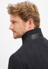 Dunkler Wollmantel mit strukturiertem Muster, ausgestattet mit einem hohen Kragen und einem schwarzen Lederakzent. Das Design ist maßgeschneidert und hat eine elegante Verarbeitung.