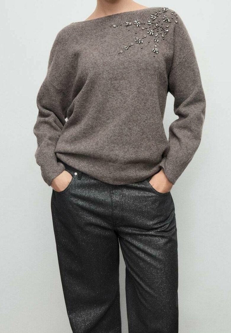 Personne portant un pull gris avec des perles ornant l'épaule et un pantalon noir scintillant, les mains dans les poches.