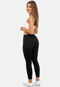 Mujer joven con cabello largo y castaño, vestida con un top corto blanco, jeans ajustados negros y zapatillas blancas, de pie y sonriendo de lado.