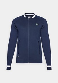 Casaco azul-marinho com fecho éclair, gola e punhos às riscas brancas e azuis, dois bolsos frontais e um pequeno logótipo bordado no peito.