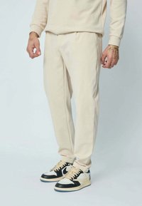 Pantalon de survêtement beige en tissu doux avec une ceinture élastique et des poches latérales. Associé à des baskets montantes noires et blanches.