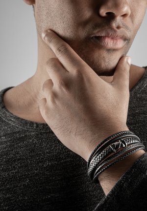 Braided black leather bracelet with silver accents, featuring a smooth, curved centerpiece. Worn on a hand resting against a chin.