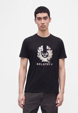 Giovane uomo che indossa una t-shirt nera Belstaff con un logo bianco consumato e jeans scuri, in piedi su uno sfondo bianco semplice.