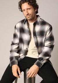 Zwart-wit geruite flanellen shirt met een kraag, knopen aan de voorkant en een borstzak; gedragen over een lichtbeige T-shirt. Zwarte spijkerbroek.