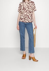 Kortärmad krämfärgad skjorta med brunt abstrakt mönster, kombinerad med blå denim, bruna klacksandaler och en vävd väska.