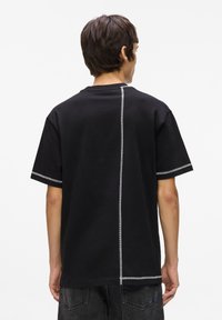 Tricou din bumbac negru cu maneci scurte, având cusături albe în contrast pe spate și la marginea manșetelor, design cu o croială lejeră.