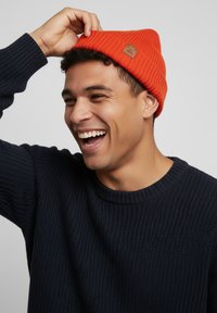 Orange knit beanie with ribbed texture and a small leather patch. Worn with a dark navy textured sweater.