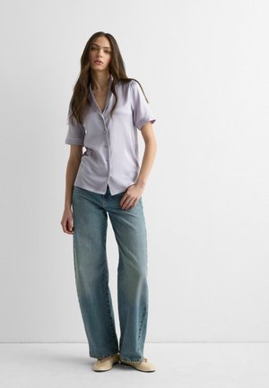 Ung kvinde med langt brunt hår iført en lys lilla kortærmet skjorte med knapper, blå jeans med vide ben og beige flade sko, stående indendørs.