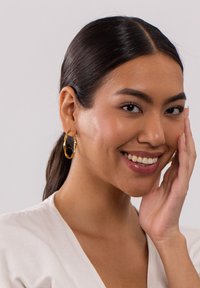 Goldene Creolen mit strukturiertem, wellenförmigem Design, die am Ohr hängen. Das Modell lächelt und berührt mit einer Hand ihr Gesicht.