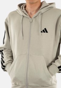 Felpa grigia chiara con zip, realizzata in un materiale morbido, dotata di una tasca frontale e strisce nere lungo le maniche con un logo nero sul petto.