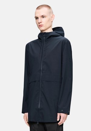 Modelo masculino con chaqueta impermeable negra con capucha, cremallera frontal, puños ajustables y un diseño minimalista sobre un fondo liso.
