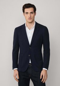 Marineblazer mit zwei Vorder pockets, zwei Knöpfen und Reverskragen, getragen über einem weißen Kragenhemd, kombiniert mit dunklen Jeans.