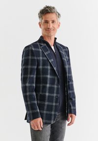 Granatowa marynarka w kratę z jasnoszarymi liniami, wykonana z teksturowanej tkaniny. Dopasowany krój z dwoma guzikami i klapami, noszona na ciemnym rozpinanym swetrze.