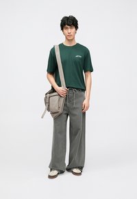 Camiseta verde de algodón, pantalones sueltos de mezclilla gris y un bolso bandolera de tela gris. El conjunto se completa con zapatos blancos.