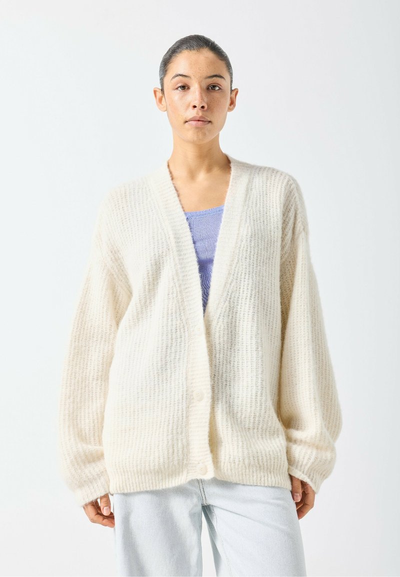 Ērts, krēmkrāsas kardigans no teksturēta dzijas auduma, ar V-veida izgriezumu, pogas slēgumu un plašām piedurknēm, valkāts virs gaiši zilas blūzes.