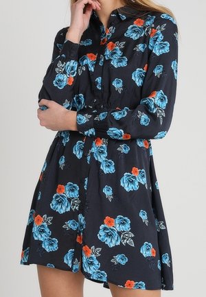 Vrouw draagt een lange mouwen zwarte jurk met blauwe en oranje bloemmotieven, staand met één hand aan haar kin en de andere over haar taille.