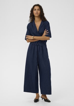 Femme aux cheveux bouclés, portant une combinaison courte à manches courtes en denim bleu foncé avec une ceinture nouée à la taille, des mules noires à talons et un bracelet en or, debout les bras croisés.