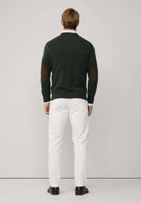 Pullover verde lavorato a maglia con toppe marroni sui gomiti, indossato sopra una camicia azzurro chiaro, abbinato a pantaloni bianchi e mocassini scuri. Vista posteriore.