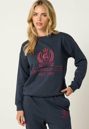 Lyshåret kvinde iført navyblå sweatshirt og bukser med rød "Equestrian Club Est. 1995" logo og hestemblem, der poserede mod en ensfarvet baggrund.