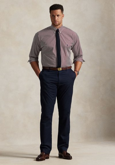 Férfi csíkos, felgörgetett hosszú ujjú ingben, sötétkék nyakkendővel, sötétkék nadrágban, barna övvel és barna bojtos mokaszinban, zsebébe dugott kézzel áll.