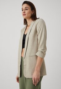 Blazer en lin beige avec revers à cran, fermeture à un bouton et manches roulées. Tissu texturé avec une coupe décontractée et une fente dans le dos.