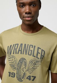 Oliivinvihreä t-paita, joka on valmistettu puuvillasta ja jossa on pyöreä kaula-aukko. Paidassa on suuri harmaa graafinen kuva pyörästä ja siivistä, jossa lukee "WRANGLER" ja "1947".