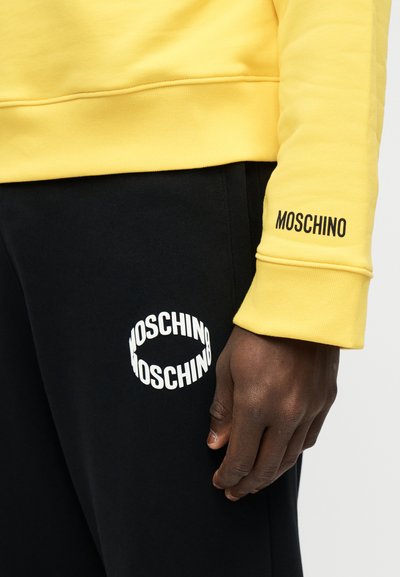 Χέρι με σκούρο δέρμα δίπλα σε μαύρο παντελόνι με λευκή επιγραφή MOSCHINO, φοράει κίτρινο φούτερ με επιγραφή MOSCHINO στη μανσέτα του μανικιού.