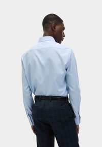 Lichtblauw overhemd met lange mouwen, geknoopte manchet en een traditionele kraag, gedragen met donker geruite broek en een zwarte riem.