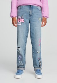 Jeans in denim azzurro chiaro con strappi frontali e stampe grafiche colorate, comprese frasi e design floreali. Abbinati a sneaker azzurro chiaro.