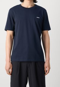 T-shirt in cotone blu navy con maniche corte, scollatura rotonda e logo bianco "HUGO" sul lato sinistro del petto. Texture liscia, vestibilità rilassata.