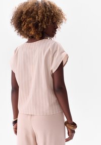 Jasnopomarańczowa bluzka z krótkim rękawem w białe pionowe paski, z okrągłym dekoltem i luźnym krojem; noszona z pasującymi jasnymi spodniami.