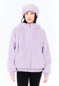 Violetti tekoturkishuppari vetoketjulla, jossa on huppu sekä ribbikuvioidut hihansuut ja vyötärö, yhdistettynä sopivaan tekoturkishattuun, valkoista taustaa vasten.