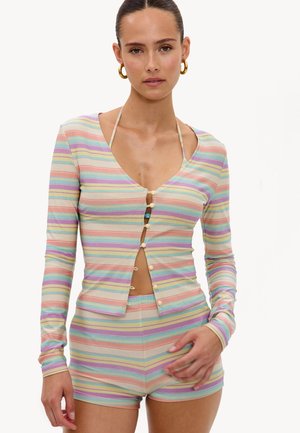 Femme portant un cardigan boutonné rayé pastel par-dessus un short assorti et un haut de bikini, avec des boucles d'oreilles anneaux en or et les cheveux tirés en arrière.