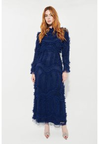 Marinblå golvlång klänning med rfflade texturer och lång ärm. Genomskinliga lager vid fållarna och dekorativa klackar med silverdetaljer.