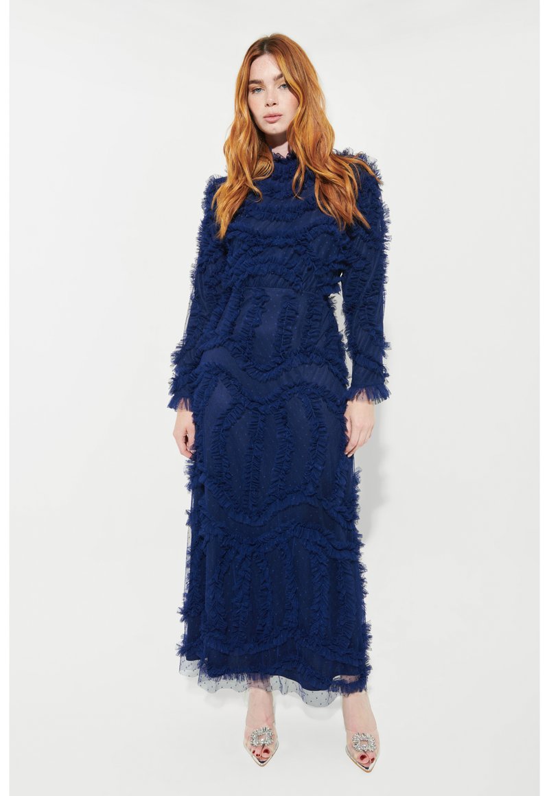 Marinblå golvlång klänning med rfflade texturer och lång ärm. Genomskinliga lager vid fållarna och dekorativa klackar med silverdetaljer.