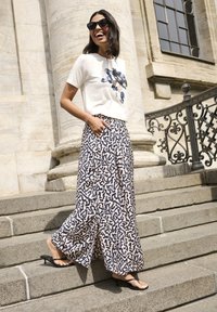 Weißes T-Shirt mit blauem Blumenmuster, kombiniert mit lockeren, gemusterten Hosen in Marineblau und Creme sowie schwarzen Flip-Flops. Treppen und Säulen im Hintergrund.