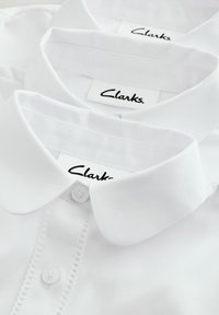 Camicie bianche a colletto di Clarks con una texture morbida. Presentano chiusure a bottone, dettagli di cucitura e etichette con il marchio "Clarks".