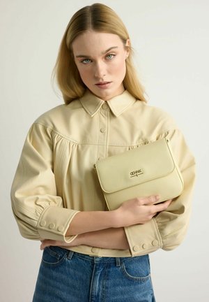 Młoda kobieta z blond włosami ubrana w beżową koszulę zapinaną na guziki i niebieskie dżinsy, trzymająca beżową torebkę na ramię z logo na przodzie.